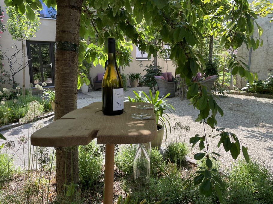 Ein Weinglas und eine Flasche stehen auf einem Holztisch im Garten. Grüne Pflanzen im Hintergrund.