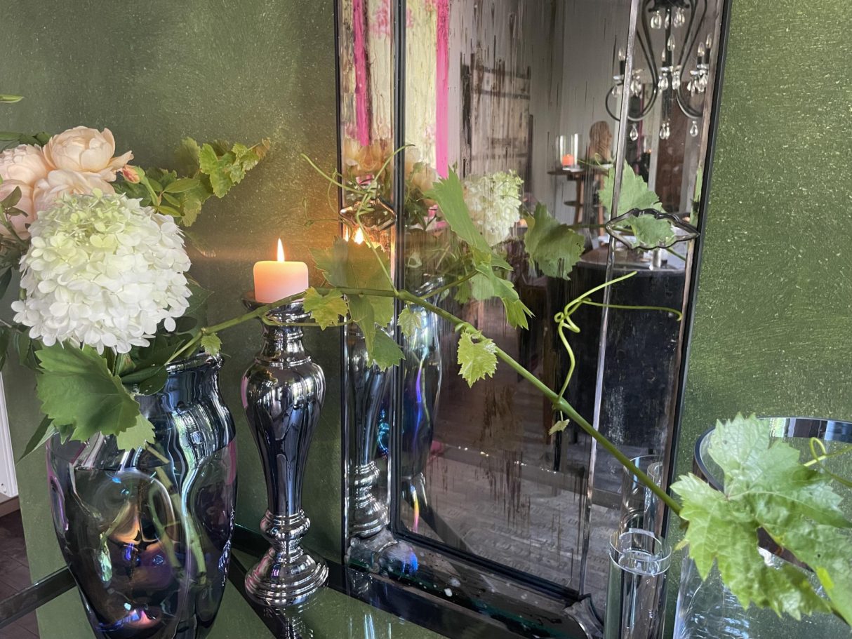 Blumenarrangement mit Kerze und Spiegel vor grüner Wand.