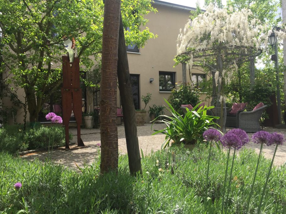 Blühender Garten mit Kunstskulptur, Bäumen und einem hellen Gebäude im Hintergrund.