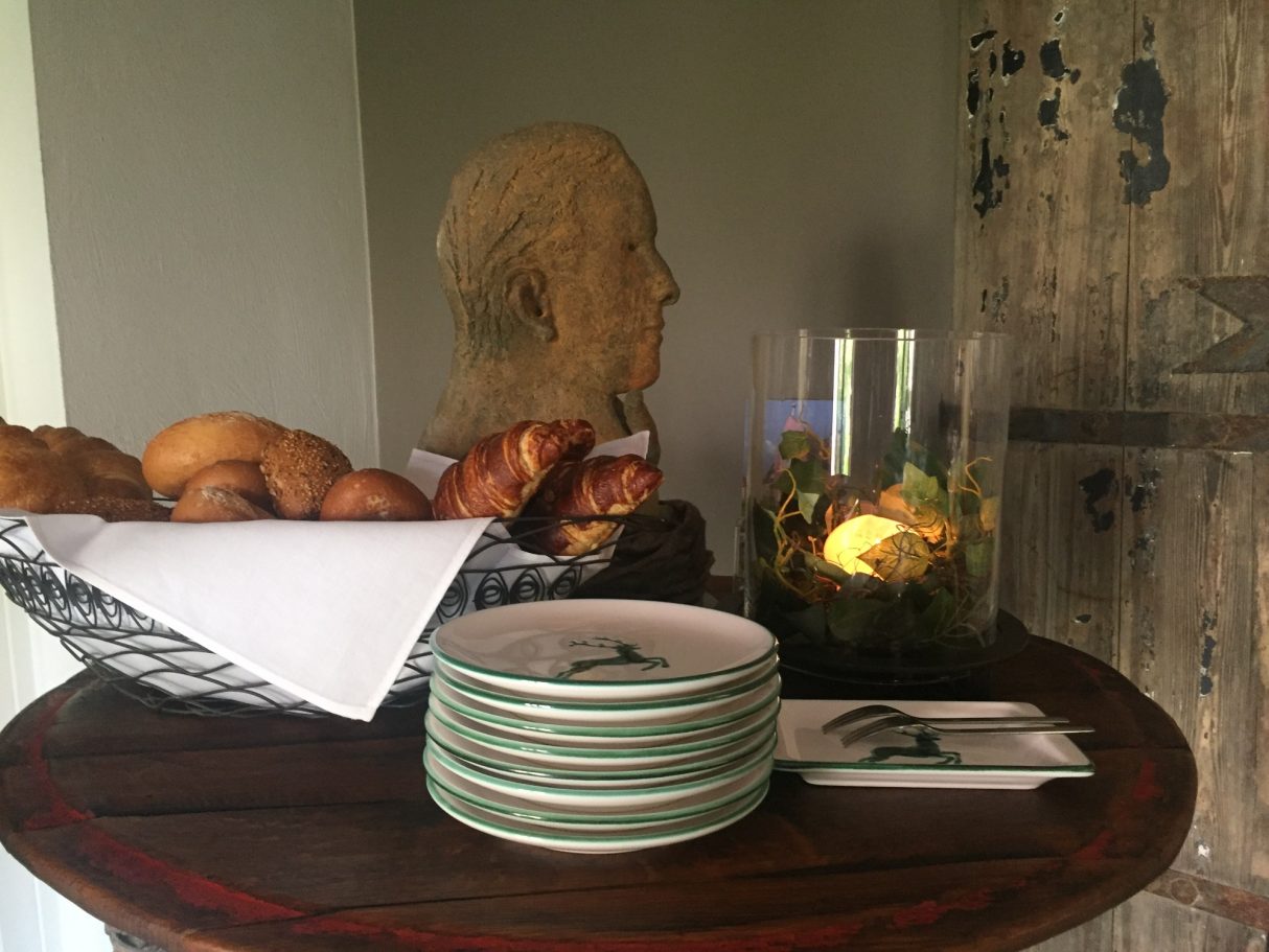 Ein Korb mit Brötchen und Croissants, daneben Teller und eine Blumenvase.