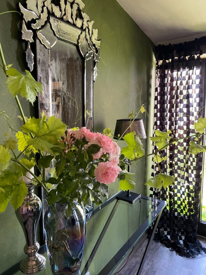 Blumenvase mit rosa Rosen, Spiegel und schwarzes Sichtschutzgitter an der Wand.