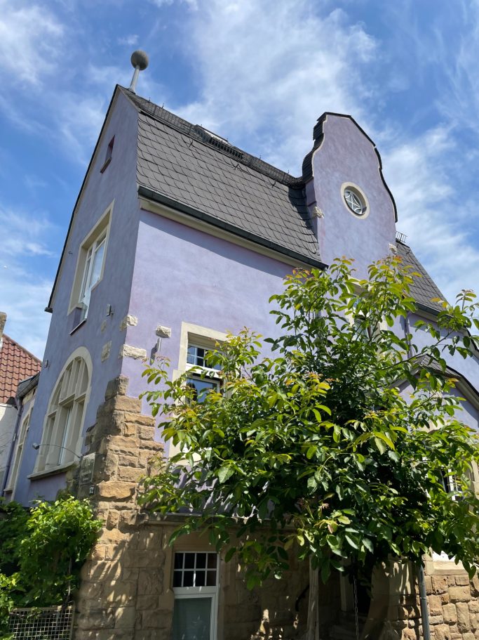 Lila Haus mit geschwungenem Dach und grünem Baum im Vordergrund.