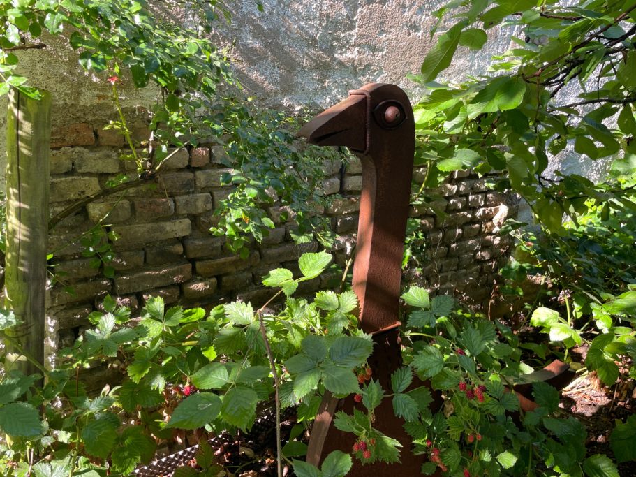 Eine stilisierte, rostbraune Vogelstatue steht umgeben von grünen Pflanzen in einem Garten.