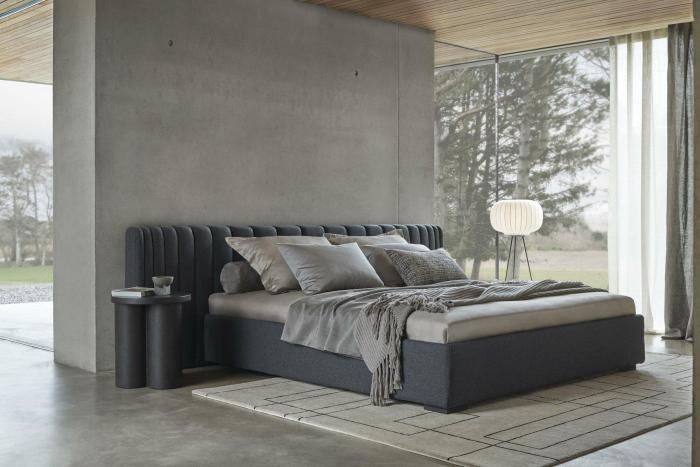 Modernes Schlafzimmer mit einem gepolsterten Bett, grauen Kissen und minimalistischem Tisch.